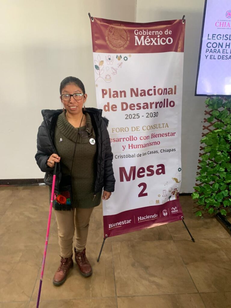 La imagen muestra a una mujer de pie junto a un cartel. Ella lleva gafas, una chaqueta negra, un suéter verde, pantalones beige y botas marrones. También sostiene un bastón blanco y rosa. El cartel a su lado tiene el logotipo del Gobierno de México y dice: "Plan Nacional de Desarrollo 2025-2030. Foro de Consulta. Desarrollo con Bienestar y Humanismo. San Cristóbal de las Casas, Chiapas. Mesa 2". En la parte inferior del cartel están los logotipos de "Bienestar" y "Hacienda". A la derecha, hay una pantalla parcialmente visible con el logotipo de Chiapas.