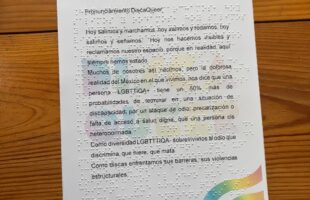 La imagen muestra un documento impreso de forma vertical sobre una hoja blanca de tamaño aproximado carta, la cual está asentada sobre una mesa de madera rústica con vetas horizontales marcadas. Se trata de un manifiesto o texto de protesta titulado "Pronunciamiento DiscaQueer". Este documento es totalmente accesible, ya que combina el texto impreso en tinta negra con el sistema de lectura táctil Braille en relieve, cuyos puntos sobresalen y son perfectamente visibles a lo largo de toda la hoja, posicionados justo encima de las líneas impresas. Detalles visuales y distribución Esquina superior izquierda: Aparece el logotipo principal con la palabra "DISCAQUEER". Cada letra del nombre está pintada con un diseño colorido que evoca la bandera del orgullo LGBTQ+, incorporando pequeños símbolos internos (como el símbolo de Venus, el de Marte y una rueda que simboliza la discapacidad motriz). Justo debajo del logo, hay una línea de texto impreso en tinta negra con letras muy pequeñas que dice: Movimiento por la accesibilidad, anticapacitista y anticuerdistas. Centro de la hoja: De fondo y de manera muy tenue (como una marca de agua gigante), se repite el logotipo "DISCAQUEER" en letras grandes y difuminadas con los colores del arcoíris. El texto principal en tinta negra se lee claramente por encima de esta marca de agua, cuyo texto dice: Título del documento: Pronunciamiento DiscaQueer Cuerpo del texto: Hoy salimos y marchamos, hoy salimos y rodamos, hoy salimos y señamos. Hoy nos hacemos visibles y reclamamos nuestro espacio, porque en realidad, aquí siempre hemos estado. Muchos de nosotros así nacimos, pero la dolorosa realidad del México en el que vivimos, nos dice que una persona LGBTTIQA+ tiene un 50% más de probabilidades de terminar en una situación de discapacidad, por un ataque de odio, precarización o falta de acceso a salud digna, que una persona cis heteronormada. Como diversidad LGBTTTIQA+ sobrevivimos al odio que discrimina, que hiere, que mata. Como discas enfrentamos sus barreras, sus violencias estructurales. (Nota: El texto en tinta se corta abruptamente en la palabra "estructurales,", dejando una coma al final, aunque los puntos en relieve Braille continúan cubriendo el resto del espacio en blanco de la hoja). Esquina inferior derecha: Se observa un elemento decorativo compuesto por tres franjas curvas y paralelas que asemejan un arcoíris ondulado o una bandera en movimiento, utilizando sutiles tonos pastel (rosa, amarillo, verde, azul y morado).