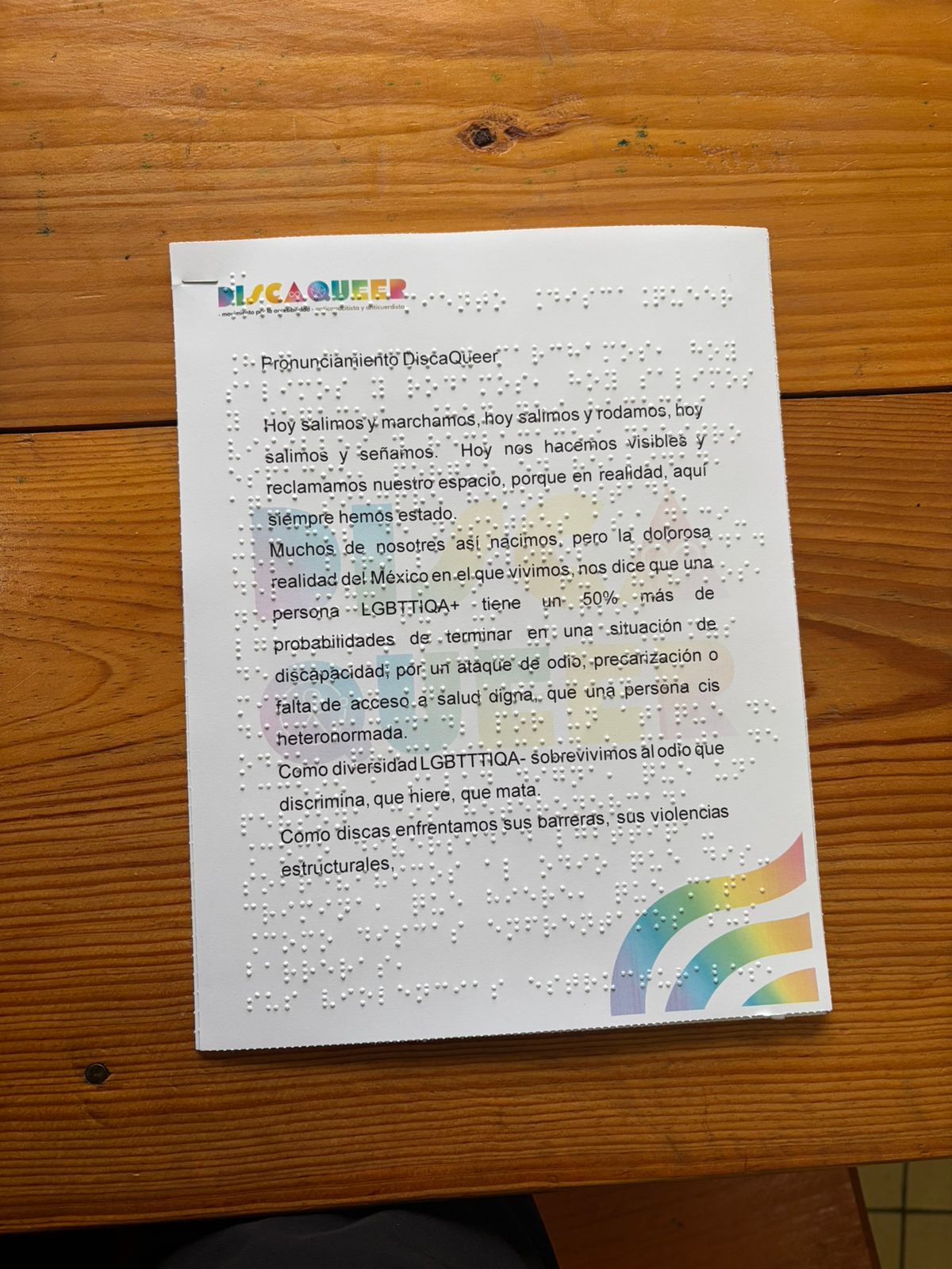 La imagen muestra un documento impreso de forma vertical sobre una hoja blanca de tamaño aproximado carta, la cual está asentada sobre una mesa de madera rústica con vetas horizontales marcadas. Se trata de un manifiesto o texto de protesta titulado "Pronunciamiento DiscaQueer". Este documento es totalmente accesible, ya que combina el texto impreso en tinta negra con el sistema de lectura táctil Braille en relieve, cuyos puntos sobresalen y son perfectamente visibles a lo largo de toda la hoja, posicionados justo encima de las líneas impresas.
Detalles visuales y distribución
Esquina superior izquierda: Aparece el logotipo principal con la palabra "DISCAQUEER". Cada letra del nombre está pintada con un diseño colorido que evoca la bandera del orgullo LGBTQ+, incorporando pequeños símbolos internos (como el símbolo de Venus, el de Marte y una rueda que simboliza la discapacidad motriz). Justo debajo del logo, hay una línea de texto impreso en tinta negra con letras muy pequeñas que dice: Movimiento por la accesibilidad, anticapacitista y anticuerdistas.
Centro de la hoja: De fondo y de manera muy tenue (como una marca de agua gigante), se repite el logotipo "DISCAQUEER" en letras grandes y difuminadas con los colores del arcoíris. El texto principal en tinta negra se lee claramente por encima de esta marca de agua, cuyo texto dice: Título del documento:
Pronunciamiento DiscaQueer
Cuerpo del texto:
Hoy salimos y marchamos, hoy salimos y rodamos, hoy salimos y señamos. Hoy nos hacemos visibles y reclamamos nuestro espacio, porque en realidad, aquí siempre hemos estado.
Muchos de nosotros así nacimos, pero la dolorosa realidad del México en el que vivimos, nos dice que una persona LGBTTIQA+ tiene un 50% más de probabilidades de terminar en una situación de discapacidad, por un ataque de odio, precarización o falta de acceso a salud digna, que una persona cis heteronormada.
Como diversidad LGBTTTIQA+ sobrevivimos al odio que discrimina, que hiere, que mata.
Como discas enfrentamos sus barreras, sus violencias estructurales.
(Nota: El texto en tinta se corta abruptamente en la palabra "estructurales,", dejando una coma al final, aunque los puntos en relieve Braille continúan cubriendo el resto del espacio en blanco de la hoja).
Esquina inferior derecha: Se observa un elemento decorativo compuesto por tres franjas curvas y paralelas que asemejan un arcoíris ondulado o una bandera en movimiento, utilizando sutiles tonos pastel (rosa, amarillo, verde, azul y morado).