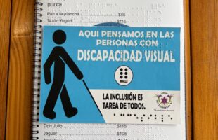 La imagen muestra el menú inclusivo con espiral negro de "Cirila" colocado de forma vertical sobre la mesa de madera rústica. La tarjeta informativa de color azul claro de la asociación "Amor sin fronteras A.C." está colocada directamente encima del menú, en la parte central, cubriendo la mayoría de las opciones de platillos, pero dejando ver el encabezado superior y las últimas opciones de la parte inferior. Al igual que el menú, esta tarjeta complementaria también cuenta con el sistema Braille en relieve superpuesto en su superficie. La tarjeta informativa central: Está dividida visualmente en dos secciones: Zona superior (Fondo azul claro): Ocupa las dos terceras partes de la tarjeta. A la izquierda muestra el pictograma negro de una persona con bastón guía. A la derecha, el mensaje en letras mayúsculas blancas y, justo debajo de este, un pequeño logotipo circular blanco que contiene un cajetín Braille de 6 puntos negros y la palabra "BRAILLE" escrita abajo. Los puntos Braille en relieve sobresalen notablemente en toda esta zona azul. (Transcripción literal) A continuación se detalla el texto visible en tinta, separando lo que pertenece a la tarjeta informativa y lo que queda al descubierto del menú: 1. Texto en la tarjeta informativa central Frase principal (Zona azul): AQUÍ PENSAMOS EN LAS PERSONAS CON DISCAPACIDAD VISUAL Detalle del símbolo (Zona azul): BRAILLE Frase secundaria (Zona blanca inferior izquierda): LA INCLUSIÓN ES TAREA DE TODOS. Logotipo de la asociación (Zona blanca inferior derecha): AMOR SIN FRONTERAS A.C. "Un desafío desde otra mirada" 2. Texto visible del menú de fondo Parte superior (Encabezado y sección Dulce): CIRILA DESAYUNOS (Todo el día) DULCE • Pan a la plancha.....................................$85 • Tazón Yogurt.........................................$115 (Nota: Las siguientes líneas de la sección dulce y la sección completa de huevos están cubiertas por la tarjeta). Parte inferior (Sección Homelets): • Don Julio................................................$115 • Jaguar....................................................$105 • Arrachera...............................................$145 • Coleto....................................................$155