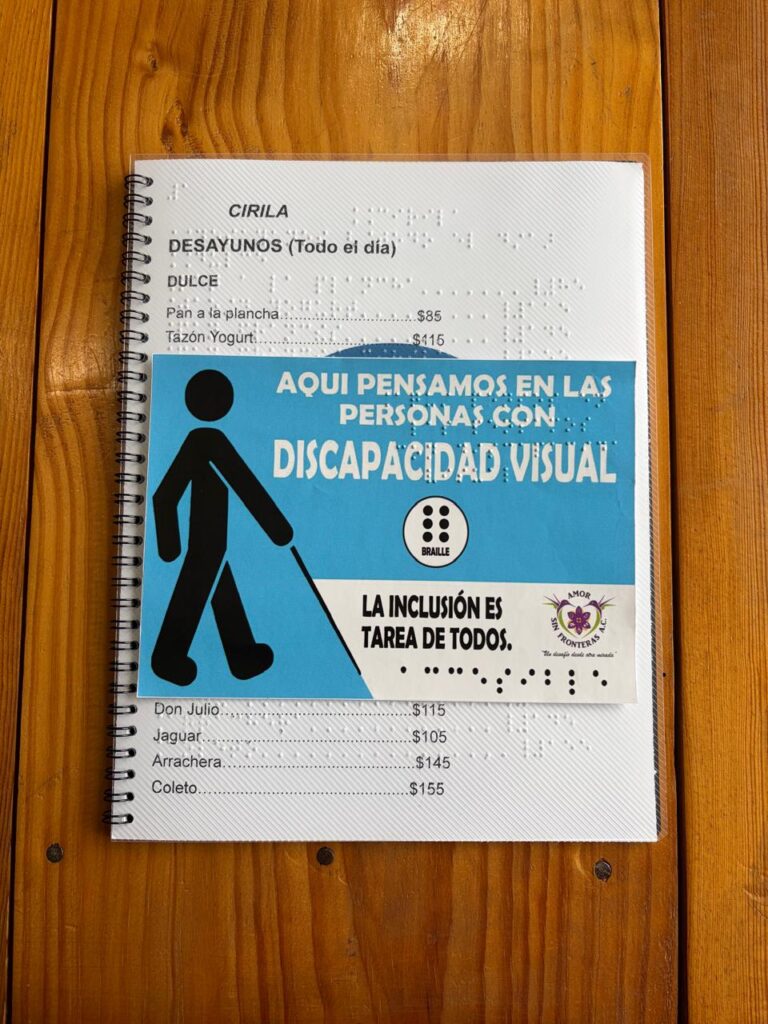 La imagen muestra el menú inclusivo con espiral negro de "Cirila" colocado de forma vertical sobre la mesa de madera rústica. La tarjeta informativa de color azul claro de la asociación "Amor sin fronteras A.C." está colocada directamente encima del menú, en la parte central, cubriendo la mayoría de las opciones de platillos, pero dejando ver el encabezado superior y las últimas opciones de la parte inferior. Al igual que el menú, esta tarjeta complementaria también cuenta con el sistema Braille en relieve superpuesto en su superficie. La tarjeta informativa central: Está dividida visualmente en dos secciones: Zona superior (Fondo azul claro): Ocupa las dos terceras partes de la tarjeta. A la izquierda muestra el pictograma negro de una persona con bastón guía. A la derecha, el mensaje en letras mayúsculas blancas y, justo debajo de este, un pequeño logotipo circular blanco que contiene un cajetín Braille de 6 puntos negros y la palabra "BRAILLE" escrita abajo. Los puntos Braille en relieve sobresalen notablemente en toda esta zona azul. (Transcripción literal) A continuación se detalla el texto visible en tinta, separando lo que pertenece a la tarjeta informativa y lo que queda al descubierto del menú: 1. Texto en la tarjeta informativa central Frase principal (Zona azul): AQUÍ PENSAMOS EN LAS PERSONAS CON DISCAPACIDAD VISUAL Detalle del símbolo (Zona azul): BRAILLE Frase secundaria (Zona blanca inferior izquierda): LA INCLUSIÓN ES TAREA DE TODOS. Logotipo de la asociación (Zona blanca inferior derecha): AMOR SIN FRONTERAS A.C. "Un desafío desde otra mirada" 2. Texto visible del menú de fondo Parte superior (Encabezado y sección Dulce): CIRILA DESAYUNOS (Todo el día) DULCE • Pan a la plancha.....................................$85 • Tazón Yogurt.........................................$115 (Nota: Las siguientes líneas de la sección dulce y la sección completa de huevos están cubiertas por la tarjeta). Parte inferior (Sección Homelets): • Don Julio................................................$115 • Jaguar....................................................$105 • Arrachera...............................................$145 • Coleto....................................................$155